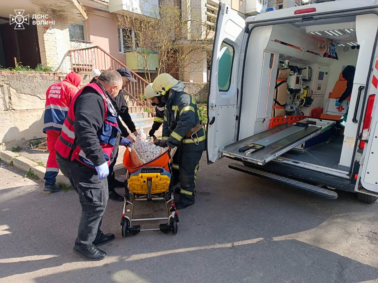 рятувальники виносять жінку, яка сама не може рухатися, до машини швидкої допомоги